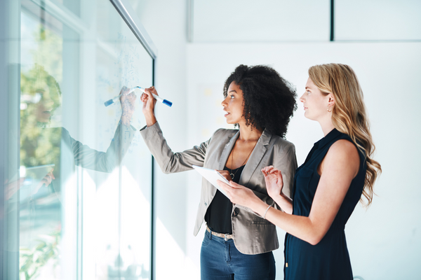 Attract a different type of audience - two women, one black, one white looking at a see through wipe board. Planning and working